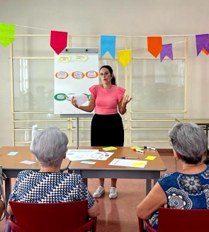 Persona de pie frente a una pizarra explicando un tema a dos personas sentadas en una mesa, con papeles y rotuladores. El fondo está decorado con banderines de colores.