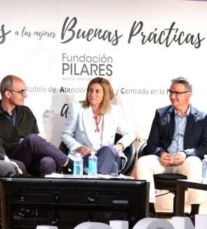Tres personas sentadas en un escenario durante un evento, con una mesa que tiene botellas de agua y micrófonos. Al fondo se ve un cartel con el texto 'Fundación Pilares' y 'Buenas Prácticas'